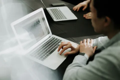 Person working on a laptop at a black table with another laptop nearby and hands visible in the background