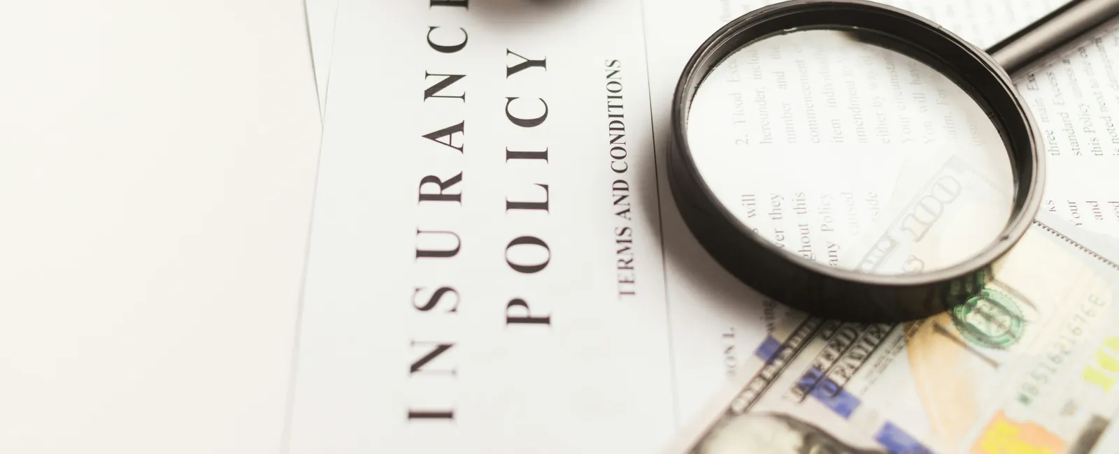 Close-up of insurance policy document with toy car, magnifying glass, and hundred-dollar bill on white surface