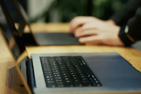 Close-up of a laptop keyboard on a wooden desk with hands typing in the background, blurred focus.