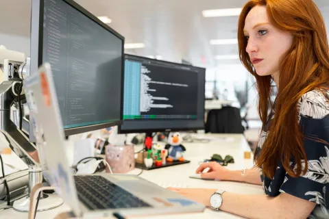 Focused woman with red hair coding on dual monitors and laptop at a modern office desk.
