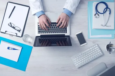 Doctor typing on laptop surrounded by stethoscope, clipboard, smartphone, keyboard, and medical documents on desk.