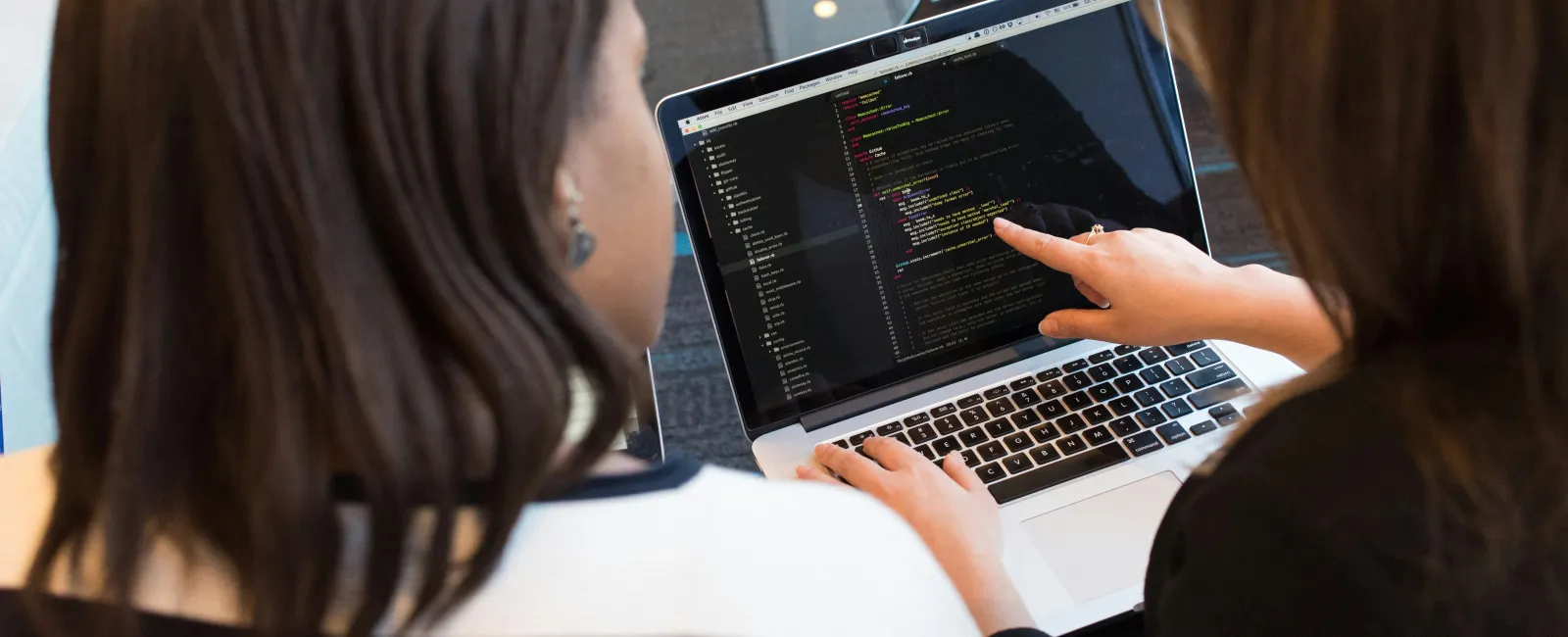 Two women collaborate and review code on a laptop screen in a modern office setting.