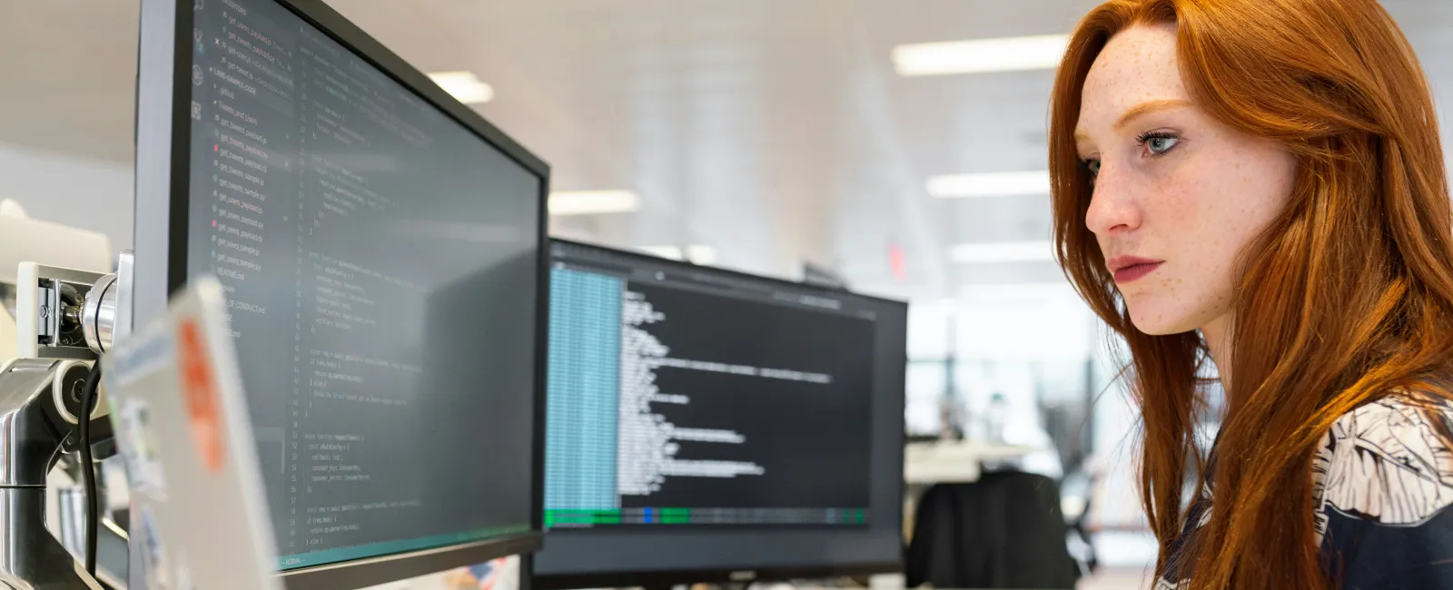 Focused woman coder working on programming with dual monitors and laptop in office environment.