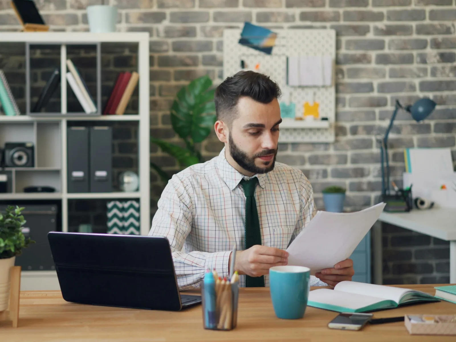 Man in office reviewing documents at desk with laptop, coffee mug, and plants in modern workspace.